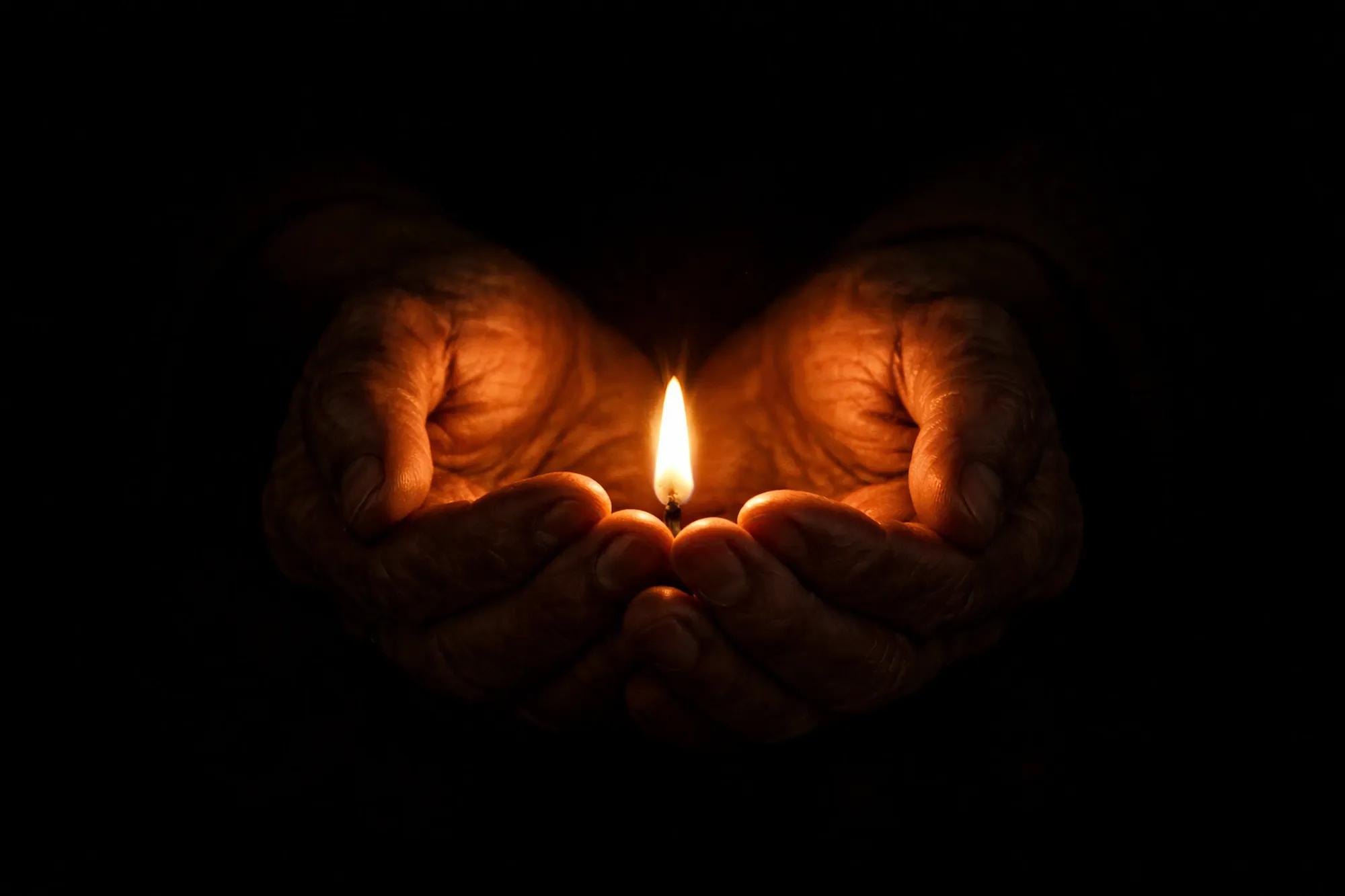 Weathered hands gently cupping a small flame in darkness, warm amber light illuminating the hands from below