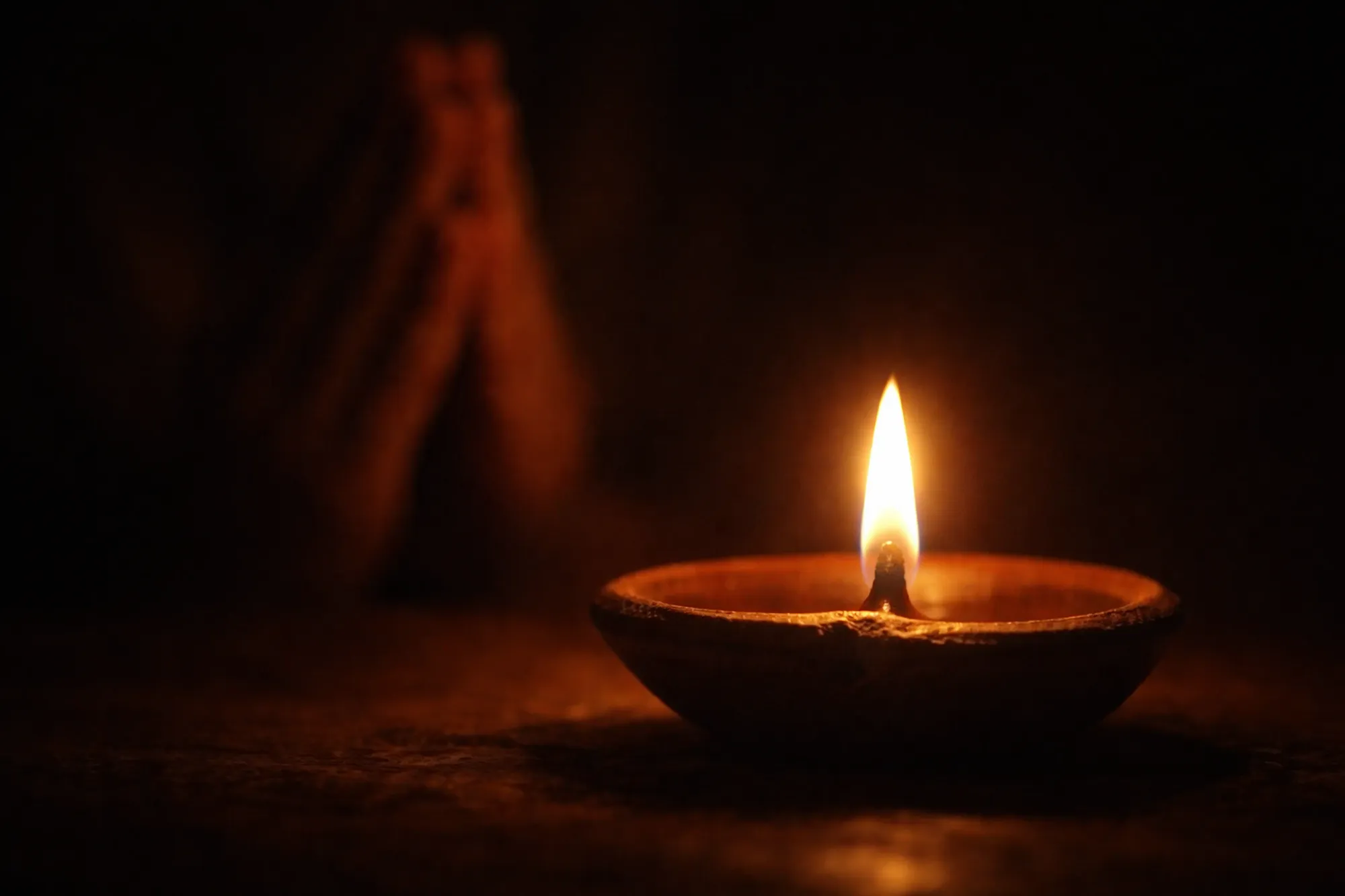 A single diya flame burning steadily in intimate darkness, folded hands barely visible in the warm golden light beside it
