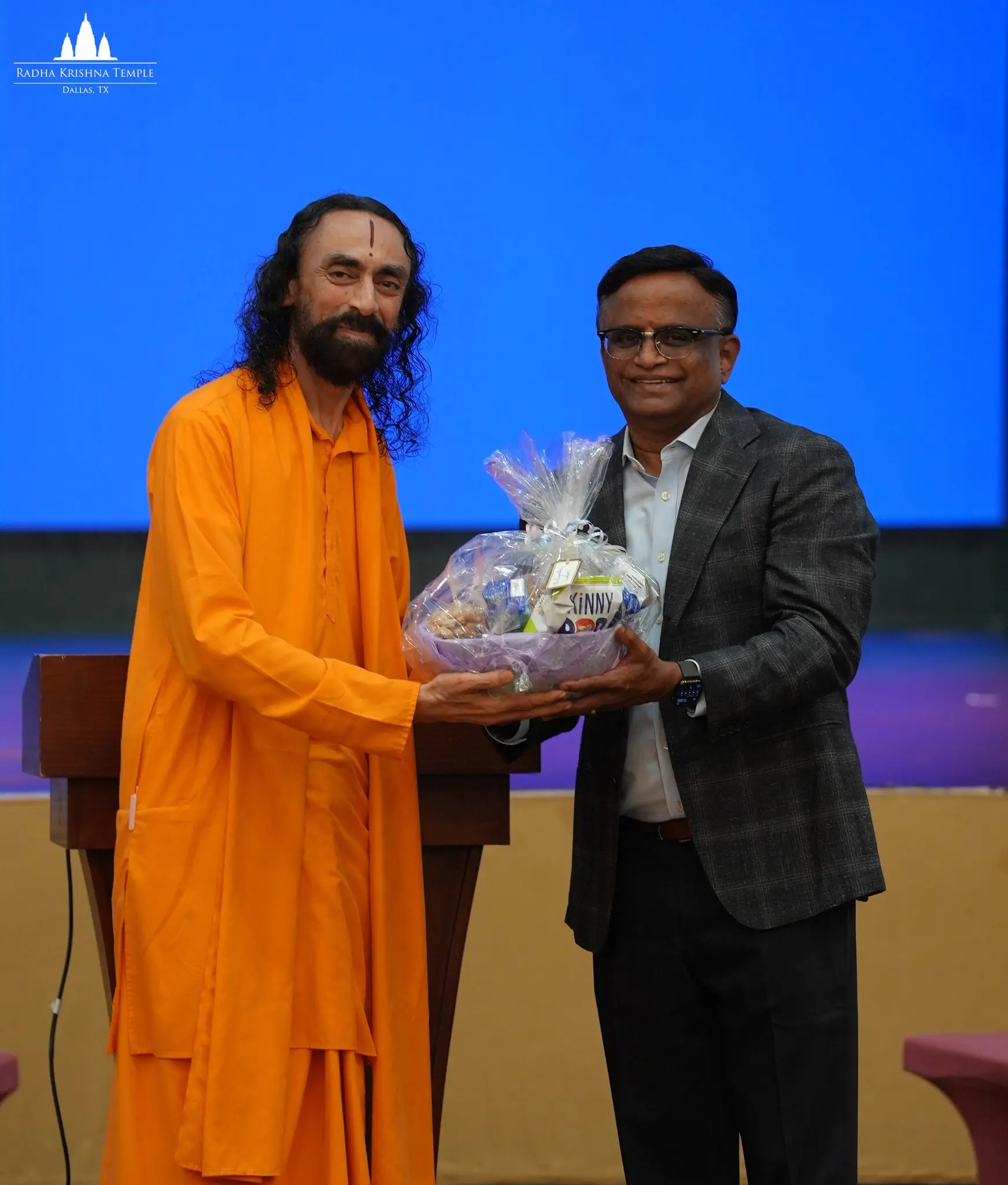 Swami Mukundananda in saffron robes receives a gift basket from a workshop organizer on stage during the Youth Leadership Workshop.