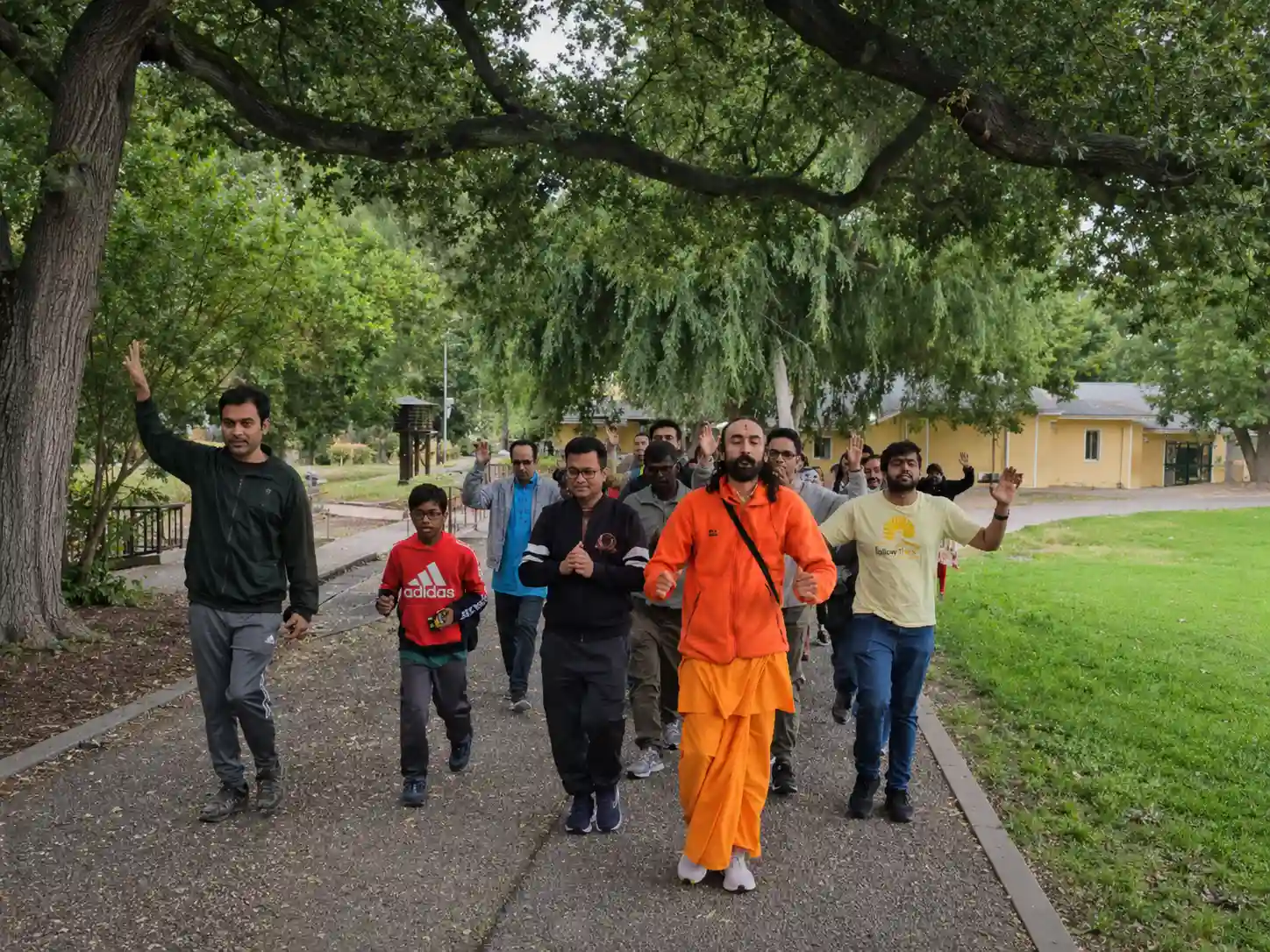 Swami Mukundananda walking outdoors with a group of participants on a tree-lined path during a morning walk.
