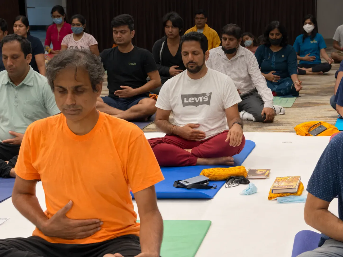 Group of participants seated on yoga mats with eyes closed, practicing meditation together in a quiet indoor hall.