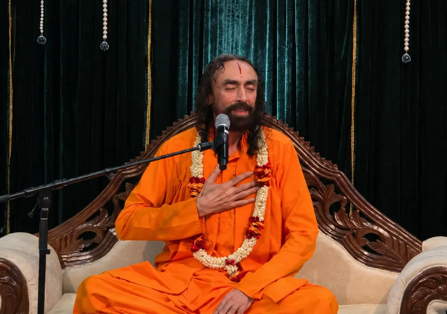 Swami Mukundananda in saffron robes seated on a carved sofa, speaking into a microphone during a spiritual discourse