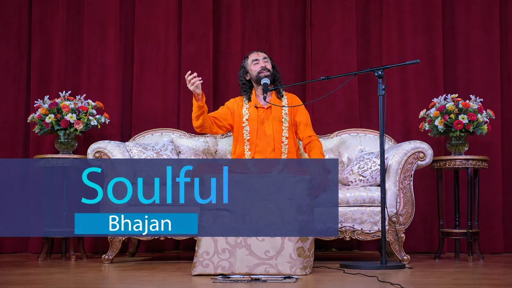 Swami Mukundananda singing a soulful bhajan on stage during the Bay Area Life Transformation Program 2026, seated before a red curtain with floral arrangements.