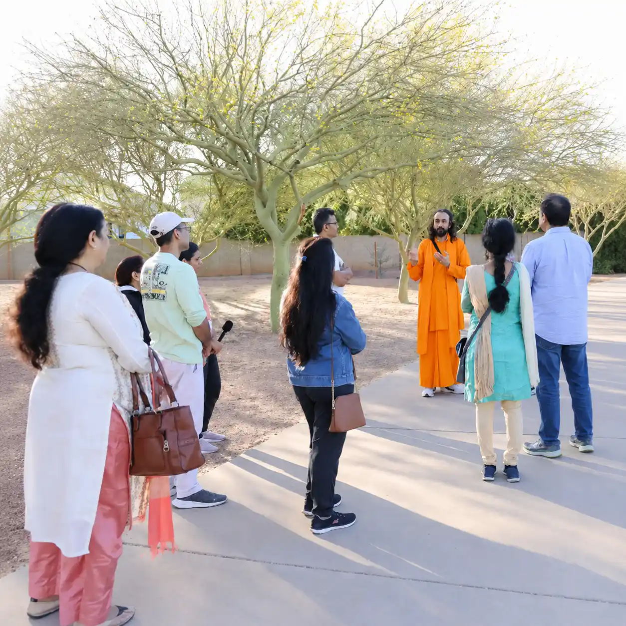 Swami Mukundananda stands outdoors in saffron robes under desert trees, speaking to a small group of devotees during the Phoenix Life Transformation Program 2026 morning walk.