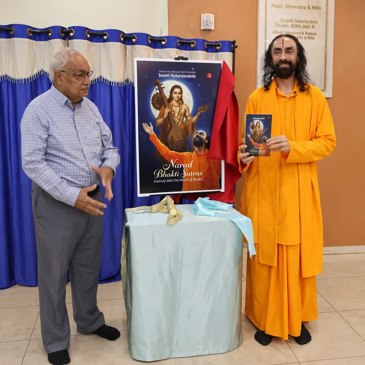 Swami Mukundananda in saffron robes holds the Narad Bhakti Sutras book beside a large poster display, with a devotee standing nearby during the unveiling event.