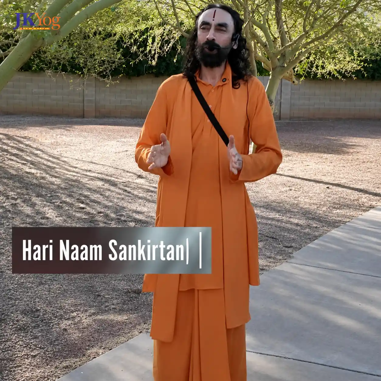 Swami Mukundananda stands outdoors in saffron robes under a tree, speaking during Hari Naam Sankirtan with a JKYog logo and text overlay on the image.