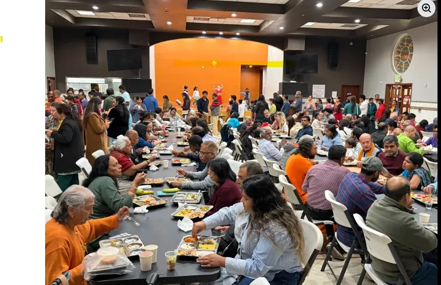 Large group of attendees seated at long tables, enjoying dinner prasadam together in an indoor hall during a community spiritual event