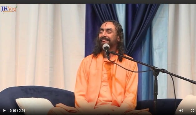 Swami Mukundananda in saffron robes seated on stage, smiling while speaking into a microphone during a spiritual talk.