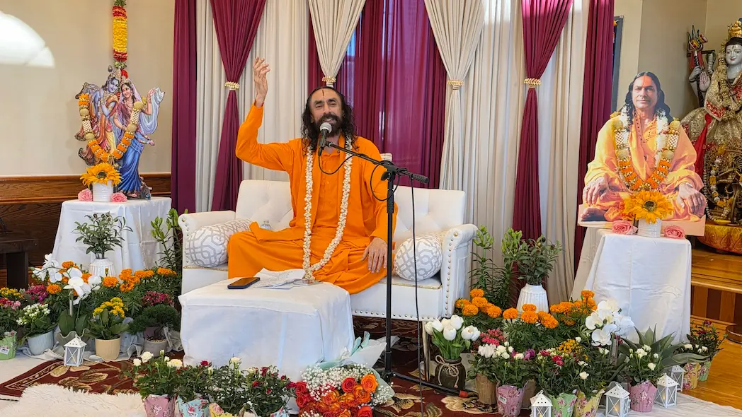 Swami Mukundananda delivering a spiritual discourse surrounded by flowers at Shree Radha Krishna Mandir.