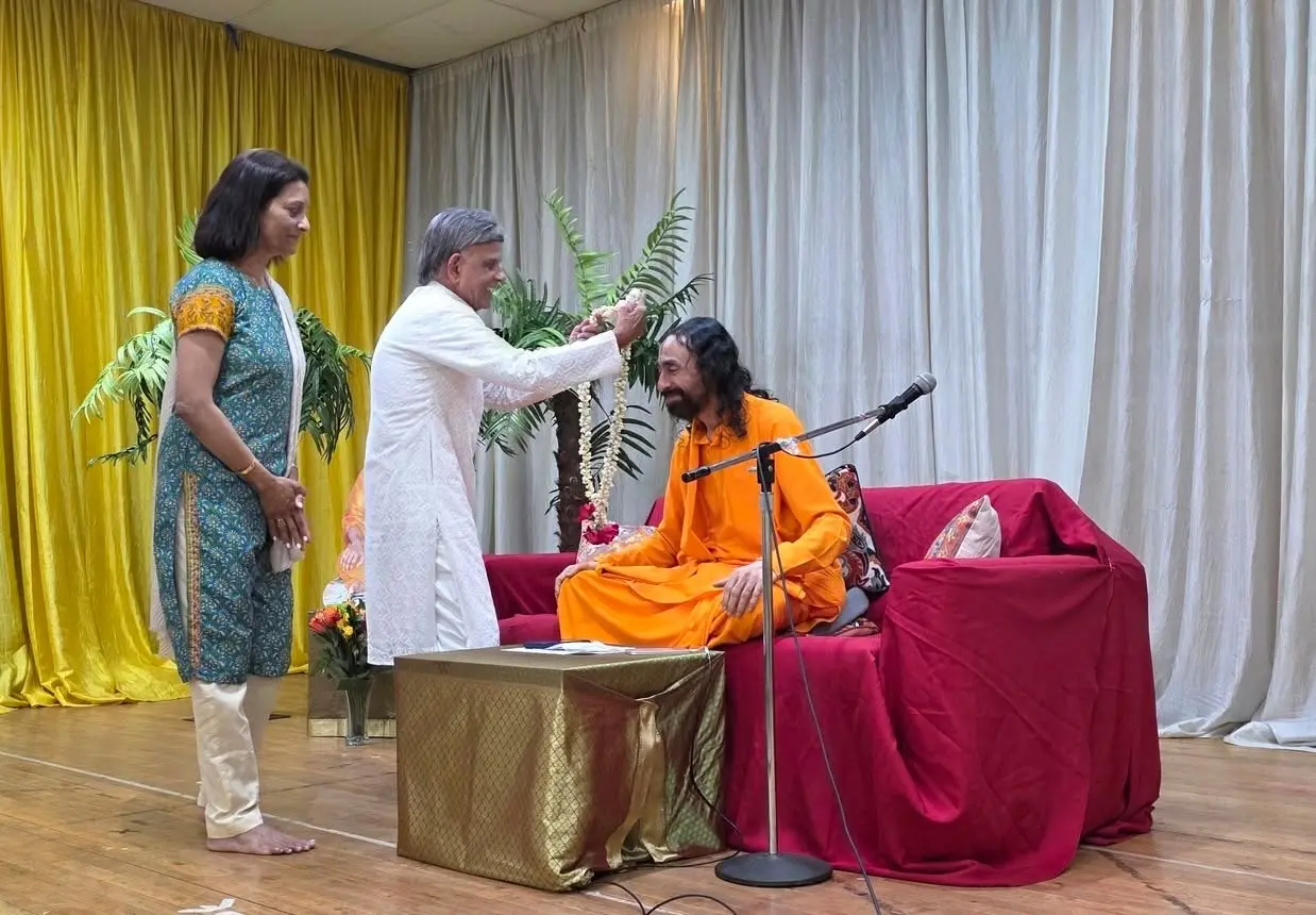 A devotee offers a garland to Swamiji seated on a stage, while another devotee stands nearby.