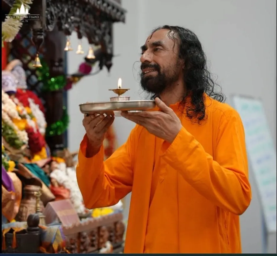 A spiritual teacher in saffron robes holds an aarti plate with a lit lamp while standing before a decorated temple altar